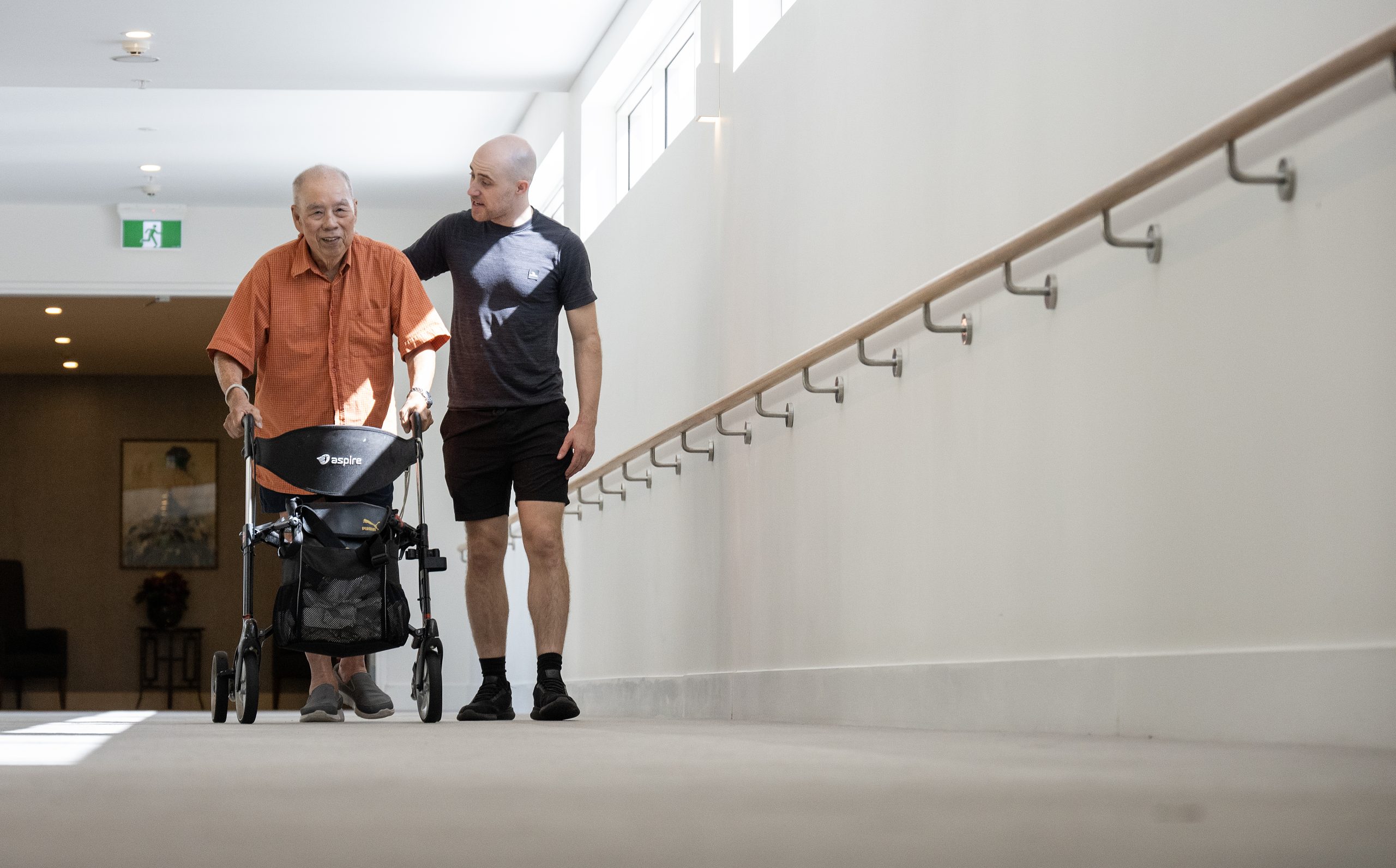 Mobile physiotherapist walking alongside an older man using a rollator in Coombabah