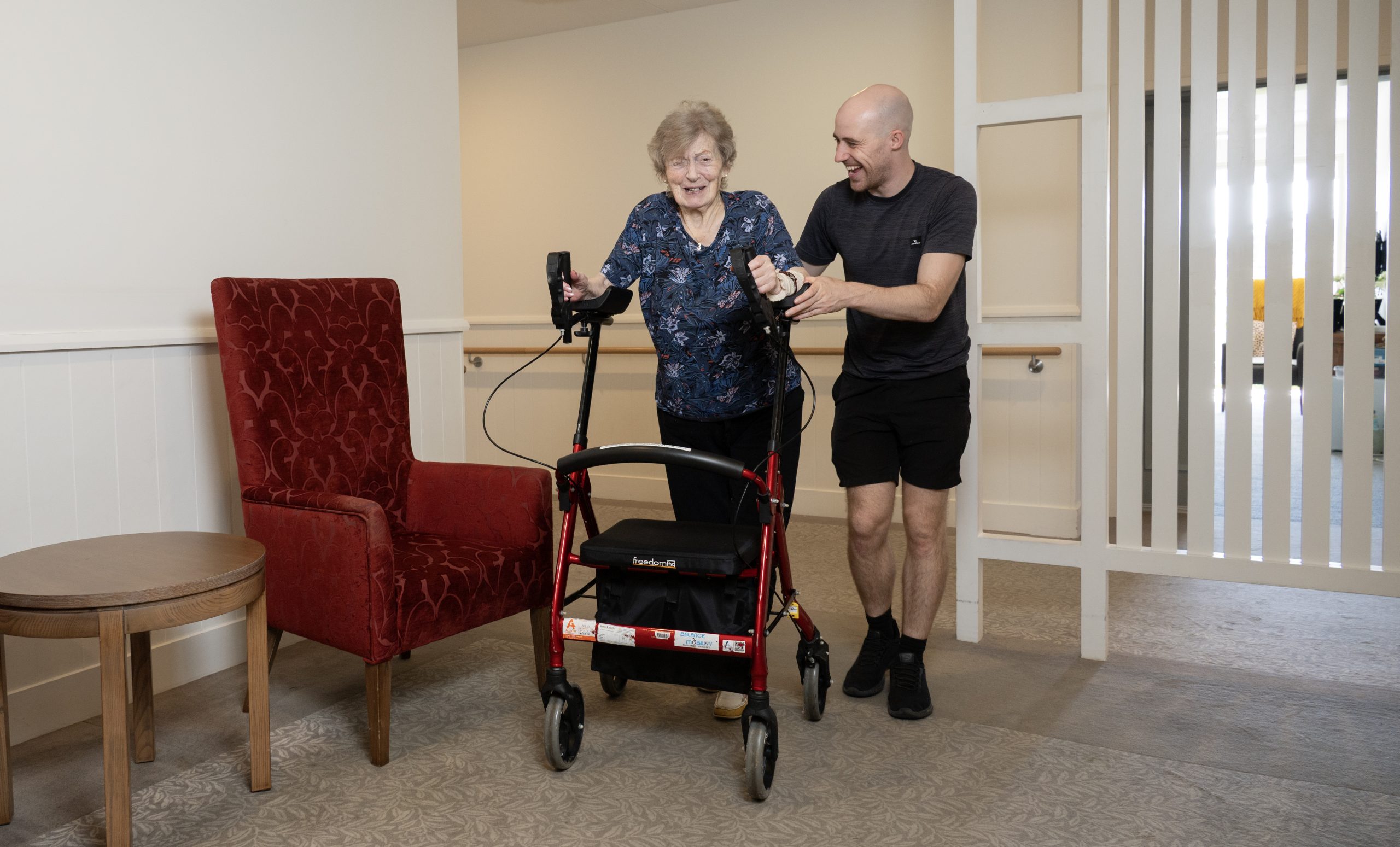 Mobile physiotherapist helping an older woman walk with a rollator during a home visit in Carrara